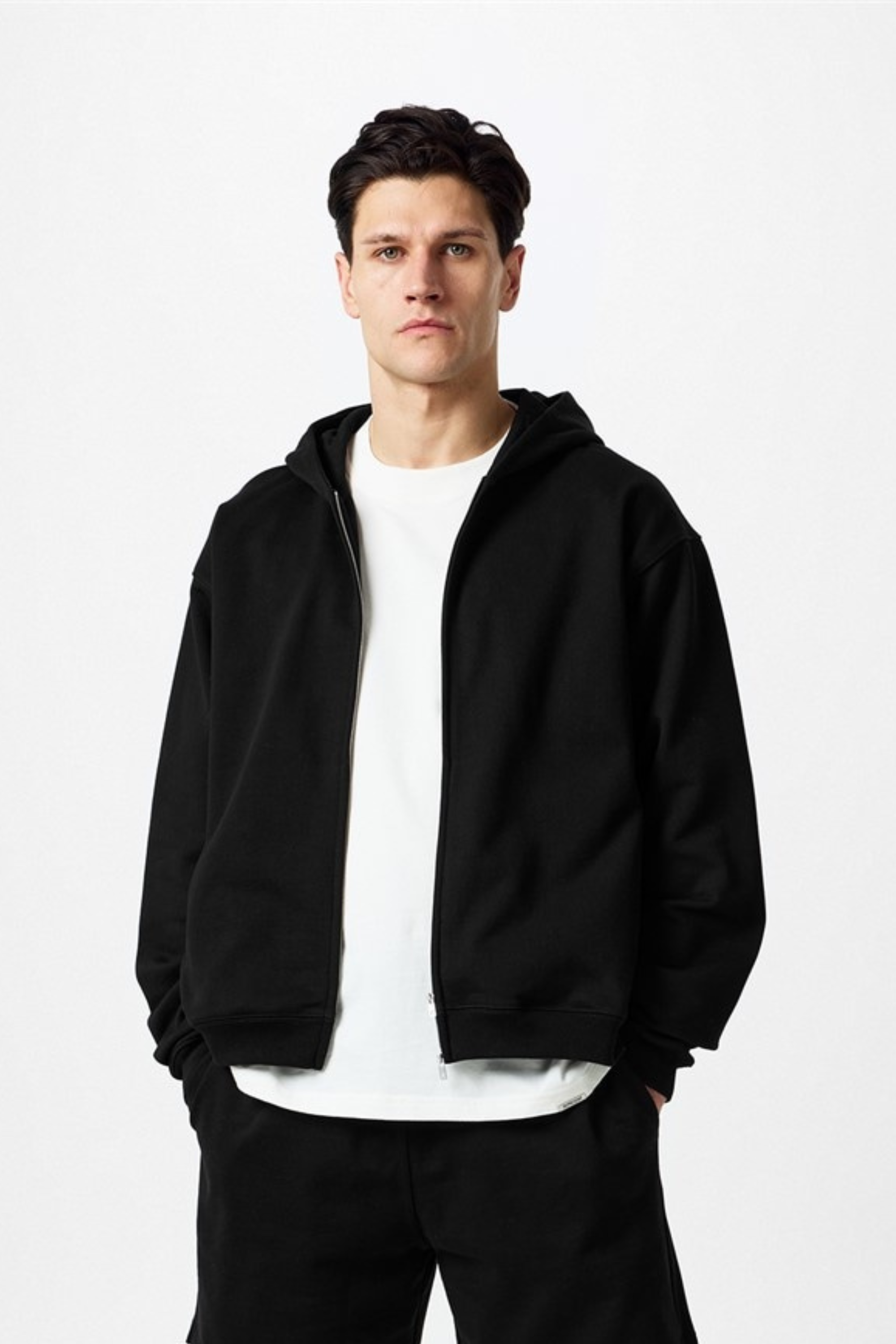 Man in black hoodie and matching pants over white t-shirt, standing against a white background.
